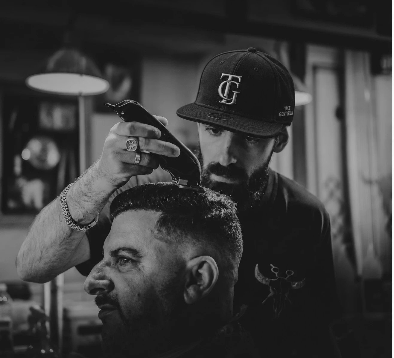 Barber v kšiltovce True Gentleman stříhá zákazníka, černobílá fotografie
